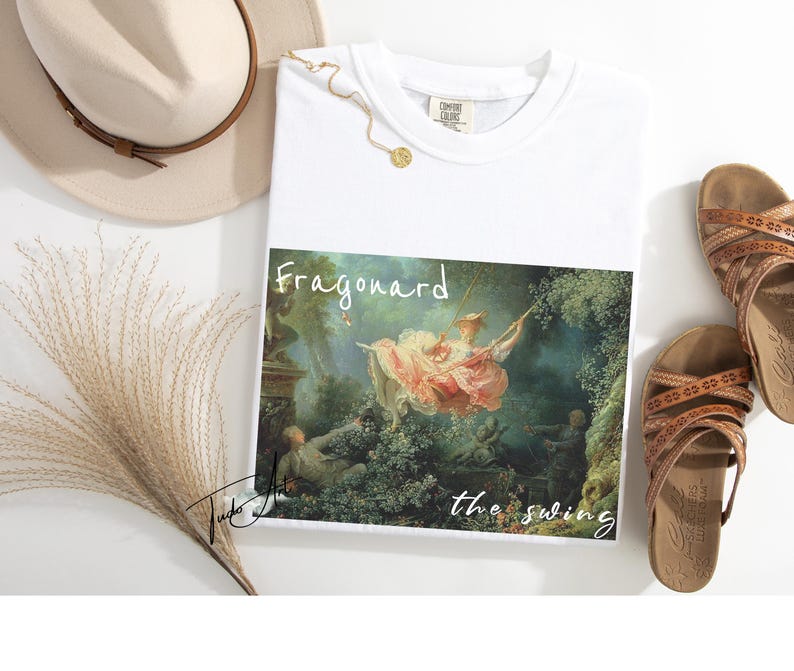 May include: White t-shirt featuring a print of Fragonard's "The Swing" painting. The shirt is paired with brown sandals, a beige hat, and a gold necklace. The artwork depicts a woman on a swing in a garden setting.