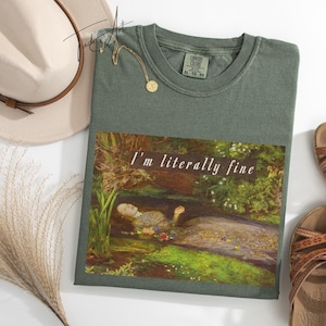 May include: Sage green t-shirt with the text "I'm literally fine" over a painting of a person in water. A beige hat, gold necklace, and brown sandals are also in the image.