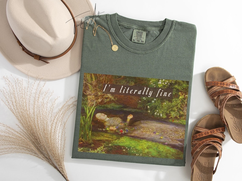May include: Sage green t-shirt with the text "I'm literally fine" over a painting of a person in water. A beige hat, gold necklace, and brown sandals are also in the image.
