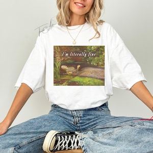 May include: White t-shirt with a rectangular graphic featuring a painting of a pond scene and the text "I'm literally fine". The shirt is worn by a person in blue jeans.
