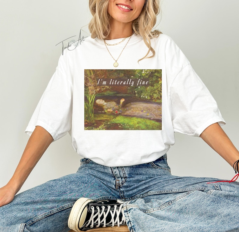 May include: White t-shirt with a rectangular graphic featuring a painting of a pond scene and the text "I'm literally fine". The shirt is worn by a person in blue jeans.