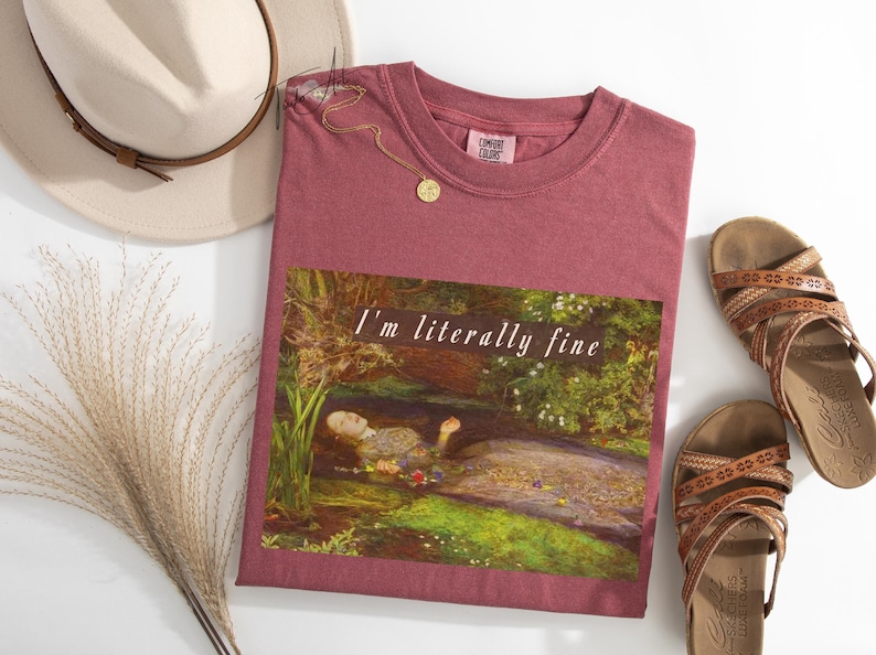 May include: A maroon t-shirt with the text "I'm literally fine" over a painting of a woman in water. The image is styled with a beige hat, gold necklace, dried grass, and brown sandals.