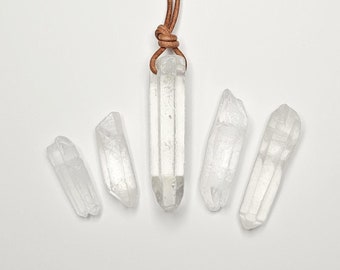 May include: A clear quartz crystal pendant suspended from a brown leather cord, accompanied by four additional loose, raw clear quartz crystal points arranged in a semi-circular pattern below the pendant on a plain white surface.