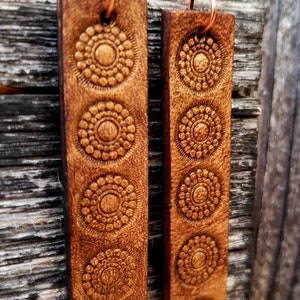 May include: A pair of brown leather earrings with a geometric pattern. The earrings are rectangular and have a copper wire hook.