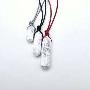 May include: Three white and gray stone pendant necklaces with different colored cords: gray, black, and red. The pendants are elongated, with a pointed bottom. The necklaces are displayed against a white background.