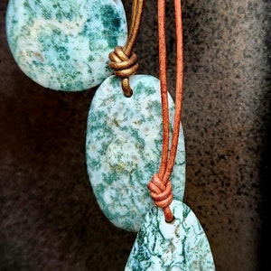 May include: Three polished moss agate pendants in teardrop, oval, and triangular shapes, each suspended from a thin, tan leather cord. The stones feature a light green base with intricate, dark green, moss-like dendritic inclusions throughout.