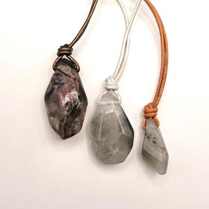 May include: Three gemstone pendants with leather and cord necklaces. The gemstones are various shapes and colors, including smoky quartz and green quartz. The cords are brown, silver, and tan. The pendants are displayed against a white background.
