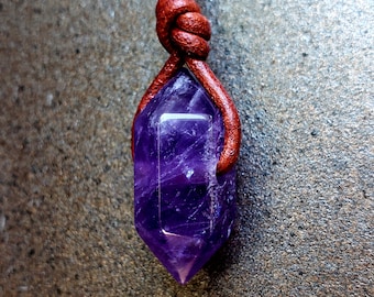 Könnte beinhalten: Ein facettierter, violetter Amethyst-Kristallanhänger, der an einer geknoteten, rotbraunen Lederschnur hängt. Der Kristall hat eine tiefe violette Farbe und eine spitze Spitze. Die Schnur ist oben geknotet.