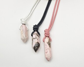 May include: Three gemstone pendant necklaces with different colored cords. The gemstones are pink and beige with dark brown inclusions. The cords are white, black, and pink. The necklaces are displayed on a white background.