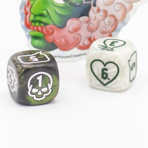 May include: A green and white sticker featuring a stylized face with a green head and red accents, with two dice. One die is black and green with a skull, the other is white with a green heart.