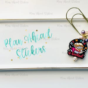 May include: A white rectangular tray with gold trim, featuring the text "Plan Ahead Stickers" in teal script. A gold paperclip holds a charm with the words "My Weekend is Booked" and decorative beads.