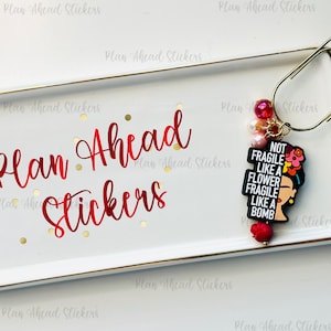 May include: A white rectangular tray with gold trim, featuring red text that reads "Plan Ahead Stickers." A gold paperclip holds a charm with a Frida Kahlo-inspired design and the words "NOT FRAGILE LIKE A FLOWER FRAGILE LIKE A BOMB."
