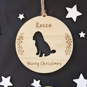 May include: A round wooden ornament with a silhouette of a dog and the text "Rocco Merry Christmas". The ornament is hanging from a string against a black background with white stars.