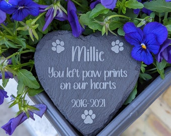 May include: A heart-shaped slate stone memorial with the name Millie engraved on it. The stone also has the text "You left paw prints on our hearts 2016-2021" and paw prints engraved on it. The stone is surrounded by purple flowers.