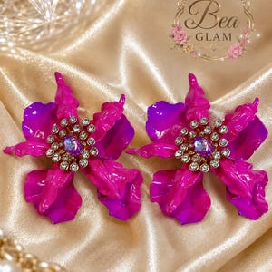 May include: A pair of flower-shaped earrings featuring vibrant fuchsia and purple petals with a central cluster of clear rhinestones surrounding a single iridescent purple stone. The text Bea GLAM is visible in the upper right corner of the image.