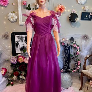 May include: A formal, floor-length, plum-colored gown with a sweetheart neckline and sheer, ruffled sleeves. The dress is displayed on a mannequin. The background includes floral decorations and a framed picture.