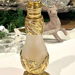 May include: A decorative perfume bottle with a frosted glass body and ornate gold-colored metal accents. The bottle features a faceted crystal stopper and leaf-like designs. The base is a gold-colored, geometric shape.