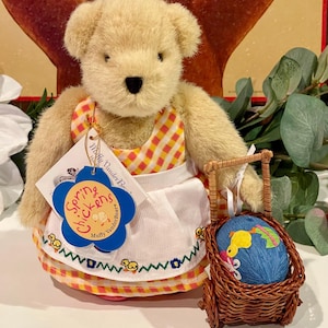 May include: A plush teddy bear wearing a yellow and orange checkered dress and white apron with a floral and wavy trim. The bear holds a small brown wicker basket with a blue egg-shaped ornament. The tag reads "Spring Chickens".