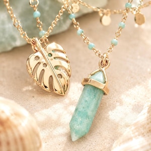 May include: Two gold-colored necklaces with pendants. One pendant is a stylized leaf, and the other is a light blue crystal. The necklaces have delicate gold chains with small blue beads.
