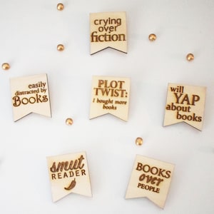 May include: Wooden banner-shaped book-themed magnets with phrases like "easily distracted by books" and "crying over fiction." The magnets are light brown with dark brown text and a small gold bead is scattered around them.