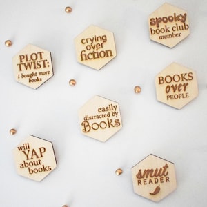 May include: Wooden hexagon-shaped book-themed magnets with engraved text. Phrases include "Plot Twist: I bought more books," "Crying over fiction," and "Spooky book club member." The magnets are light brown with dark brown text.