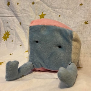 Puede incluir: Un juguete de peluche con forma de cubo de tela rosa, azul y blanca. El juguete tiene dos pequeños ojos negros y dos brazos cortos azules.