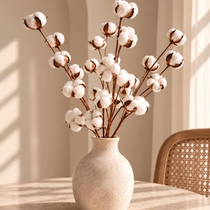 Könnte beinhalten: Eine beige Keramikvase mit strukturierter, gesprenkelter Oberfläche, die einen Strauß getrockneter Baumwollzweige enthält. Die Baumwollzweige bestehen aus mehreren flauschigen, weißen Baumwollkapseln an dünnen, braunen, holzigen Ästen.