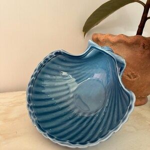 May include: A blue ceramic seashell-shaped bowl. The bowl has a glossy finish and a ribbed texture, with a scalloped edge. The bowl is sitting on a light-coloured surface.