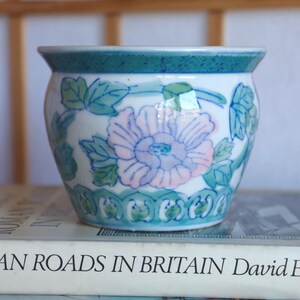 May include: A small, round ceramic planter with a floral design. The planter has a white base with a large pink and blue flower, surrounded by green leaves and vines. The rim is teal green with a decorative pattern. The planter is on top of a book.