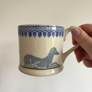 May include: A cream-coloured ceramic mug featuring a blue decorative border pattern along the top rim and a blue graphic illustration of a dog in a resting position on the side. The mug has a curved handle and a smooth, glossy finish throughout.