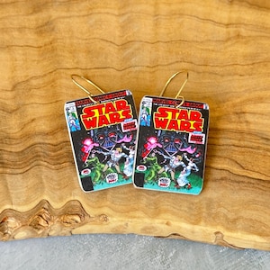 May include: A pair of Star Wars comic book earrings. Each earring features a miniature comic book cover with the title "STAR WARS" in red and white. The artwork depicts characters and Darth Vader. The earrings have gold-colored hooks.
