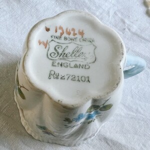 Pu&ograve; includere: Una tazza da t&egrave; bianca con un motivo floreale blu e un manico blu. Il fondo della tazza ha un'etichetta che recita "1924 Fine Bone China" e "Shelley's England R272101".