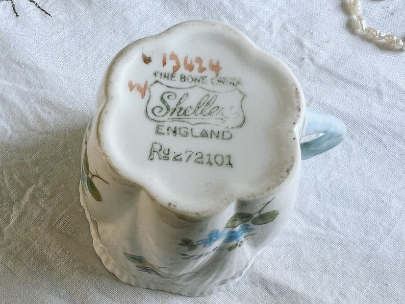 Pu&ograve; includere: Una tazza da t&egrave; bianca con un motivo floreale blu e un manico blu. Il fondo della tazza ha un'etichetta che recita "1924 Fine Bone China" e "Shelley's England R272101".