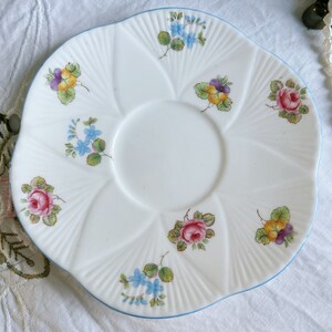 Pu&ograve; includere: Un piattino bianco con un bordo blu e disegni floreali. Il piattino ha un bordo ondulato e presenta una variet&agrave; di fiori, tra cui rose, violette e nontiscordardime.
