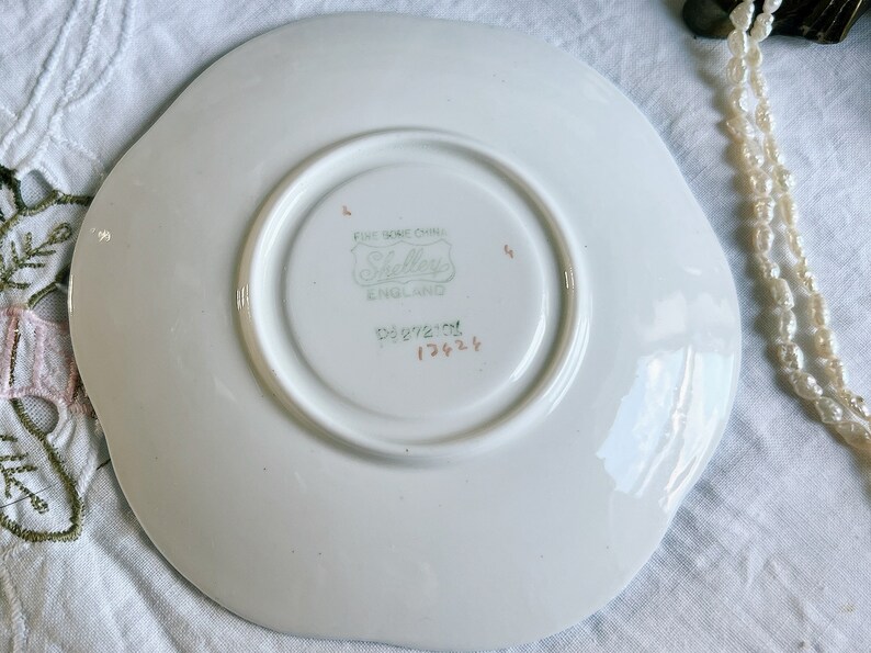 Pu&ograve; includere: Piatto bianco con bordo ondulato. Il piatto ha un logo Shelley Fine Bone China verde e bianco e il numero 13424 in marrone.
