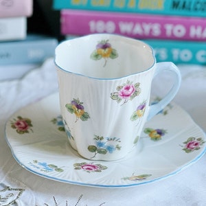 Pu&ograve; includere: Una tazza da t&egrave; e un piattino bianchi con un bordo blu e un motivo floreale. La tazza ha un delicato disegno floreale con fiori rosa, gialli e viola. Il piattino ha un motivo floreale simile con un bordo blu.