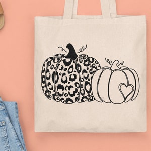 May include: A white tote bag with black handles features a design of two pumpkins. One pumpkin is a leopard print and the other is a solid pumpkin with a heart in the center.