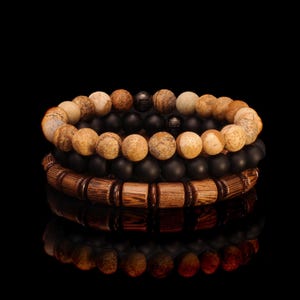 May include: Three stacked bracelets. The top two bracelets feature round, speckled, tan and black beads. The bottom bracelet has rectangular wooden beads. The background is black.