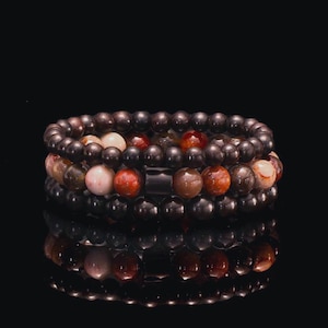 May include: Three stacked beaded bracelets. The top and bottom bracelets feature dark brown beads. The middle bracelet has a mix of red, tan, and cream-colored beads. The bracelets are displayed on a reflective black surface.
