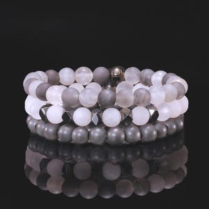 May include: Three stacked beaded bracelets in shades of gray and white. The bracelets feature round beads and geometric metal accents. The beads have a matte finish, and the bracelets are displayed against a black background.