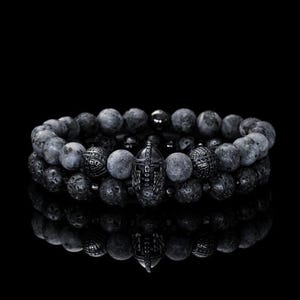 May include: Two beaded bracelets. One bracelet features dark gray, round beads with a silver-colored Spartan helmet charm. The other bracelet has textured, dark gray beads. The bracelets are displayed on a reflective black surface.