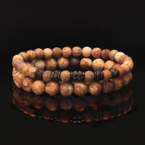 May include: Three stacked beaded bracelets. The bracelets feature a mix of round, light brown, and tan beads. Dark metal accents are interspersed throughout the design, creating a contrast. The bracelets are displayed on a reflective black surface.