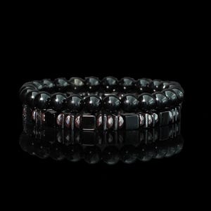 May include: Two black beaded bracelets. The top bracelet features round beads, while the bottom bracelet has a mix of square and faceted beads. The bracelets are displayed on a reflective black surface.