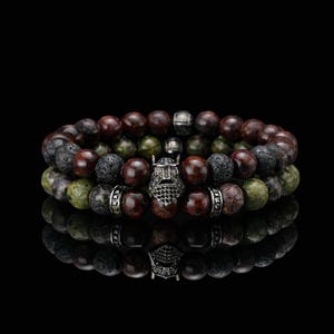 May include: Three beaded bracelets with a dark background. The bracelets feature a mix of dark red, green, and black beads. Each bracelet has a silver-colored metal accent, and one has a Viking helmet charm.