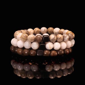 May include: Three stacked beaded bracelets. The top bracelet features light brown and tan beads. The middle bracelet has white and brown beads. The bottom bracelet is made of dark brown beads. Each bracelet has a black square bead.