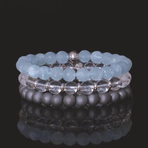 May include: Three beaded bracelets stacked on top of each other. The top bracelet has light blue beads, the middle one has clear beads, and the bottom one has dark gray beads. The bracelets are displayed on a reflective black surface.