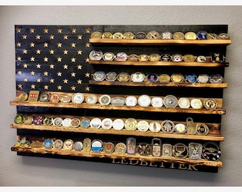 May include: A wooden American flag with five shelves for displaying challenge coins. The flag is black with white stars and has a distressed wood finish. The shelves are made of natural wood and are stained a dark brown color. The flag is mounted on a wall and has the name "LEDBETTER" engraved on the bottom.