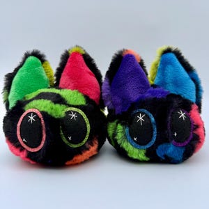 May include: Two plush cat-shaped toys with large, glittery eyes and colorful fur. One has green, pink, and orange accents, while the other features purple, blue, and pink. Both have black fur and white star details.