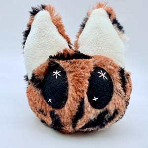 May include: A plush toy shaped like a stylized cat face. The toy features a brown and black spotted pattern, large black eyes with white star accents, and white ears with black and brown fur trim. The toy is soft and cuddly.