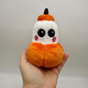 May include: A plush toy shaped like a pumpkin with a white face, orange body, and a brown stem. The toy has large black eyes with white star details and pink cheeks. The plush is soft and fluffy.